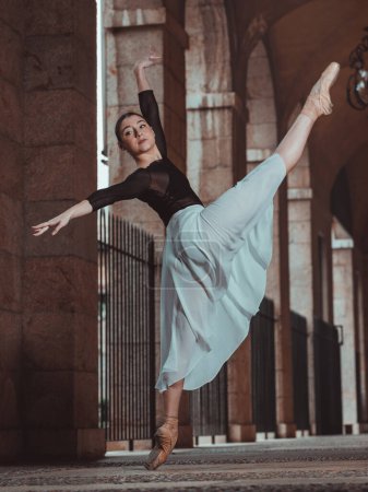 Photo for Side view of full body ballerina in silk tutu and pointe shoes raising leg and arms while performing ballet positions in arched passage of building - Royalty Free Image
