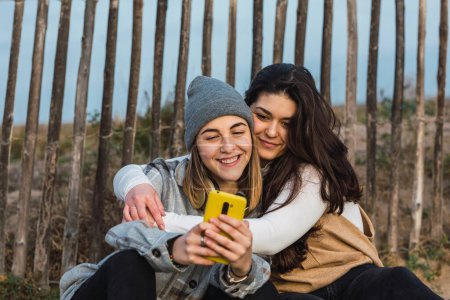 Téléchargez les photos : Jeune femme aux longs cheveux foncés dans des vêtements décontractés assis sur un chemin en bois en terrain rural et embrassant une meilleure amie à l'aide d'un smartphone - en image libre de droit