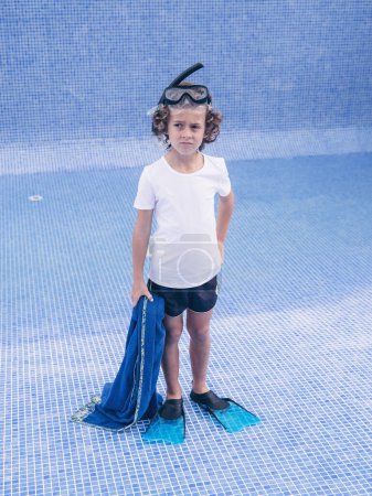 Foto de Cuerpo completo de preadolescente insatisfecho en máscara de snorkel y aletas con toalla de pie en el fondo de la piscina vacía y mirando hacia otro lado infelizmente - Imagen libre de derechos