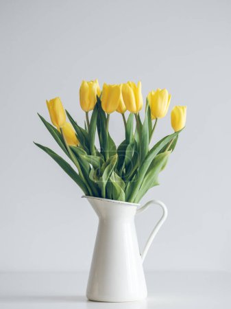 Foto de Ramillete de vivos tulipanes amarillos colocados en jarrón de cerámica sobre mesa contra pared blanca en estudio - Imagen libre de derechos