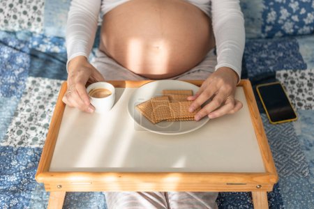 Foto de Desde arriba de la hembra embarazada suave sentado en la cama con bandeja y beber café con galletas durante el desayuno por la mañana - Imagen libre de derechos
