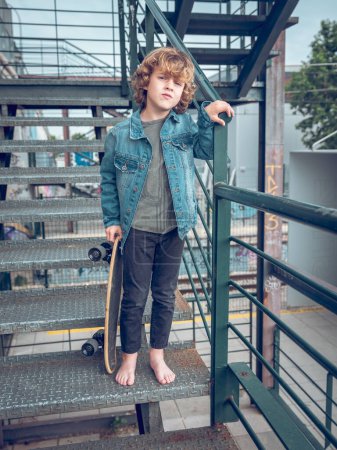 Téléchargez les photos : Corps complet de garçon mignon calme en vêtements décontractés avec penny conseil debout sur l'escalier tout en s'appuyant sur la rampe sur la rue - en image libre de droit