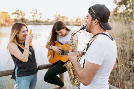 Foto de Joven hembra en traje casual bebiendo cerveza y riendo mientras mira a sus amigos tocando la guitarra y el saxofón en un día soleado cerca del lago - Imagen libre de derechos