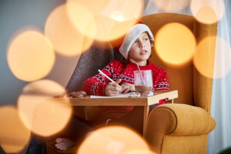 Photo for Cute boy in Santa hat looking into distance and sitting in armchair while writing letter for Santa Claus having cacao in room with bokeh during Christmas celebration - Royalty Free Image