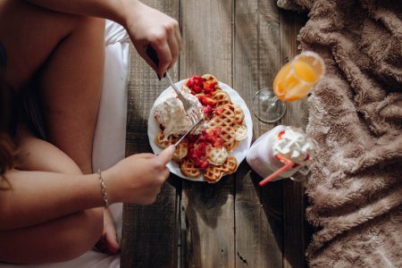 Téléchargez les photos : Jeune femme mangeant gaufres appétissantes et boissons savoureuses sur plateau au lit - en image libre de droit