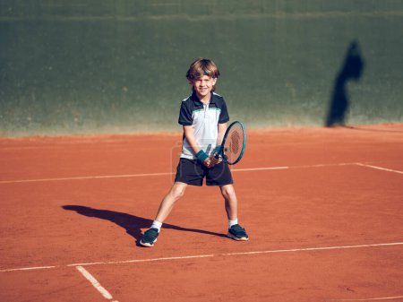 Téléchargez les photos : Pleine longueur de petit garçon sportif positif en vêtements de sport et baskets tenant la raquette tout en jouant au tennis sur le terrain pendant l'entraînement en plein air par une journée ensoleillée - en image libre de droit