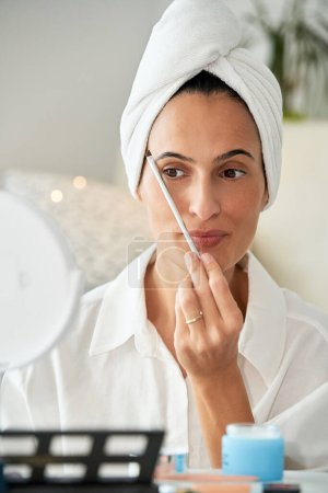 Foto de Hembra adulta enfocada con toalla en la cabeza aplicando cepillo cosmético cerca de la ceja mientras hace maquillaje en casa por la mañana y mirando el espejo redondo - Imagen libre de derechos