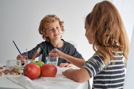 Foto de Niño positivo mirando linda chica mientras pintan juntos en la mesa con manzanas maduras en la sala de luz sobre fondo blanco - Imagen libre de derechos