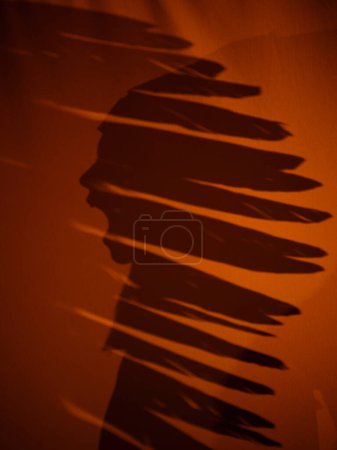 Photo for Side view silhouette of faceless boy with opened mouth in traditional native American headdress on textile on evening time in dark room - Royalty Free Image