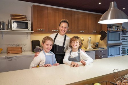 Photo for Brother and sister hugged by smiling woman wearing aprons standing at kitchen counter after cooking and looking at camera - Royalty Free Image