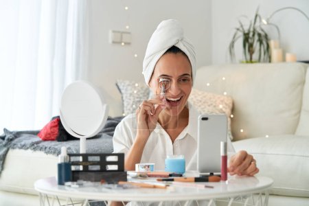 Téléchargez les photos : Délicieuse femme souriante adulte avec serviette sur la tête appliquant friseur de cils tout en faisant maquillage et diffusion blog de beauté en ligne sur smartphone à la maison - en image libre de droit