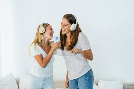Téléchargez les photos : Amis féminines positives avec smartphone moderne chantant tout en écoutant de la musique dans les écouteurs sans fil dans le salon lumineux à la maison - en image libre de droit