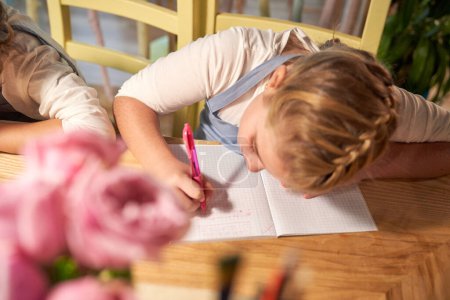 Téléchargez les photos : D'en haut de la jeune fille blonde assise à table et écrivant dans un copybook tout en faisant de l'exercice en classe - en image libre de droit