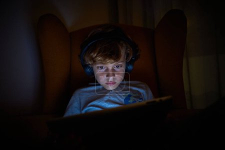 Téléchargez les photos : Garçon sérieux dans les écouteurs sans fil modernes jouant au jeu vidéo sur tablette tout en étant assis dans un fauteuil dans la pièce sombre la nuit - en image libre de droit
