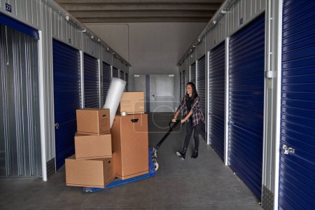 Photo for Full body side view of content female rolling manual pallet truck with carton boxed into rented container during moving day - Royalty Free Image