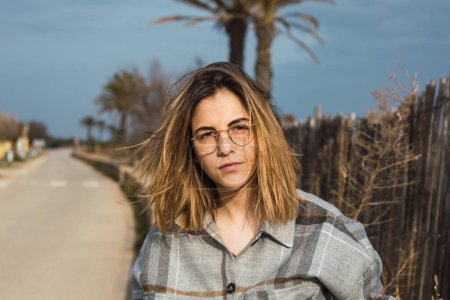 Photo for Self assured young female millennial in stylish casual clothes and eyeglasses near rustic wooden fence and looking at camera - Royalty Free Image