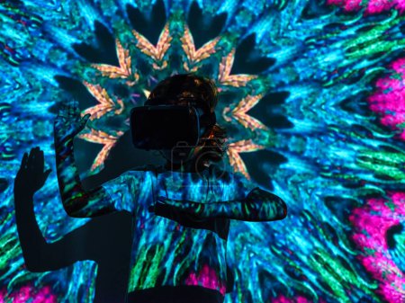 Photo for Anonymous child in VR headset playing in virtual reality while standing with raised hands under bright neon abstract colorful projector illumination - Royalty Free Image