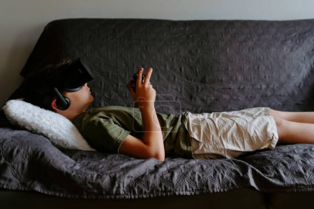 Photo for Side view of concentrated child in casual clothes lying on sofa with gamepad and enjoying VR goggles device - Royalty Free Image