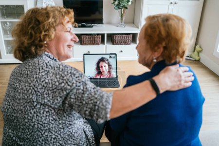 Photo for Back view of cheerful senior women chatting with friend during online video meeting via laptop while staying at home during coronavirus lockdown - Royalty Free Image