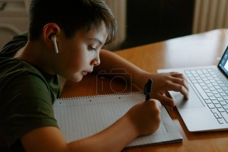 Foto de Vista lateral del niño enfocado en auriculares inalámbricos usando computadora portátil y anotando información mientras realiza la tarea - Imagen libre de derechos
