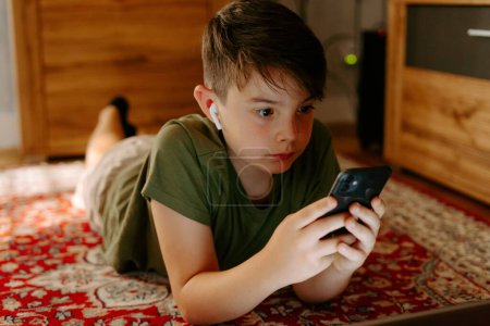 Foto de Niño concentrado en camiseta y verdaderos auriculares inalámbricos acostado en la alfombra en casa y viendo video en el teléfono inteligente durante las vacaciones de verano - Imagen libre de derechos