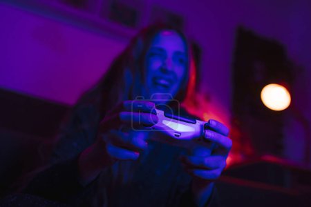 Foto de Vista de cerca de las manos de una chica sonriente sosteniendo un controlador y jugando videojuegos. Ella está en una sala de estar con luces de colores por la noche. - Imagen libre de derechos