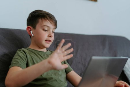 Téléchargez les photos : Petit garçon joyeux dans de vrais écouteurs sans fil agitant la main tout en bavardant avec des amis sur un ordinateur portable assis sur le canapé à la maison - en image libre de droit