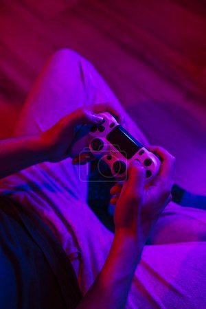Téléchargez les photos : Vue rapprochée des mains d'un garçon jouant à des jeux vidéo assis sur le sol avec des lumières colorées - en image libre de droit