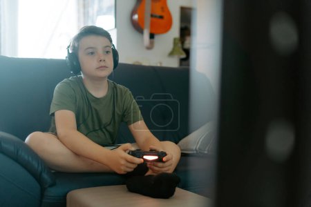 Photo for Concentrated little boy in casual clothes sitting on sofa with crossed legs and playing video game with joystick - Royalty Free Image