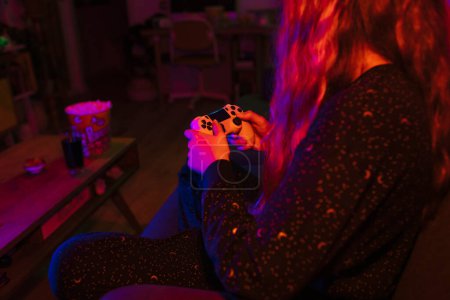 Téléchargez les photos : Vue latérale d'une fille blonde méconnaissable jouant à des jeux vidéo avec des lumières colorées dans le salon. - en image libre de droit