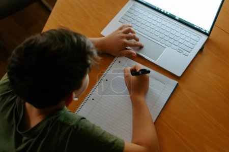 Photo for High angle of focused little boy in wireless earphones using laptop and writing down information while doing homework assignment - Royalty Free Image