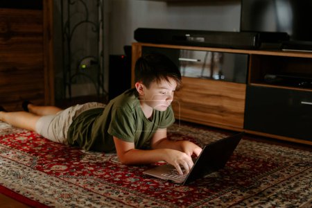 Téléchargez les photos : Pensif petit garçon en vêtements décontractés couché sur le sol ordinateur portable de navigation pendant le week-end à la maison - en image libre de droit