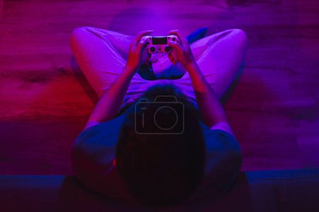 Téléchargez les photos : Vue du dessus d'un garçon assis sur le sol jouant à des jeux vidéo la nuit avec des lumières colorées - en image libre de droit