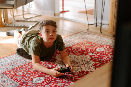 Photo for From above of little boy in comfy clothes lying on floor while playing video game with joystick at home - Royalty Free Image