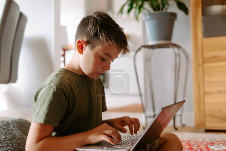 Photo for Serious little boy sitting on floor with legs crossed and typing on laptop keyboard at home - Royalty Free Image
