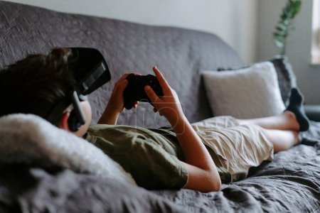 Téléchargez les photos : Enfant concentré en vêtements décontractés couché sur un canapé avec manette de jeu et bénéficiant d'un dispositif de lunettes VR - en image libre de droit
