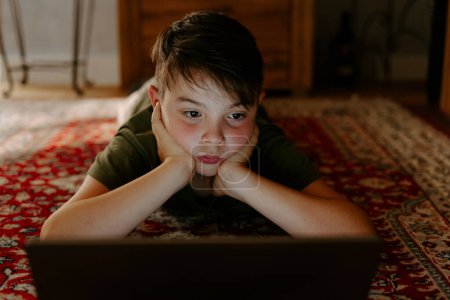 Photo for Pensive little boy in casual clothes lying on floor with hands at chin and watching movie on laptop during weekend at home - Royalty Free Image