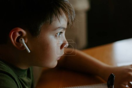 Téléchargez les photos : Vue latérale du petit garçon concentré dans les écouteurs sans fil à l'aide d'un ordinateur portable et d'écrire des informations tout en faisant des devoirs - en image libre de droit