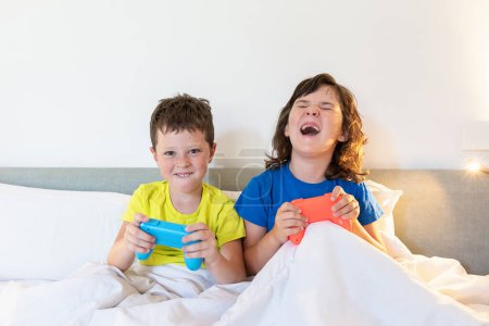 Téléchargez les photos : Contenu frères et sœurs avec des manettes de jeu jouant jeu vidéo intéressant tout en étant assis ensemble dans un lit confortable sous couverture dans la chambre à coucher à la maison - en image libre de droit