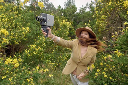 Téléchargez les photos : Femme mince souriante en tenue décontractée et chapeau flânant avec une vieille caméra vidéo près des buissons avec des fleurs en fleurs colorées à la campagne et regardant loin - en image libre de droit