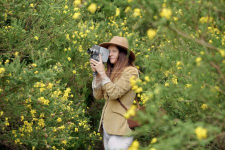 Foto de Mujer delgada sonriente en ropa casual y sombrero paseando con vieja cámara de video y estuche de cuero cerca de arbustos con flores florecientes de colores en el campo y mirando hacia otro lado - Imagen libre de derechos