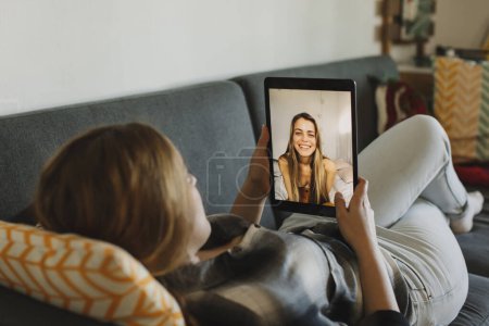 Téléchargez les photos : Vue en gros plan d'une jeune fille blonde faisant un appel vidéo avec une amie allongée sur son canapé. - en image libre de droit