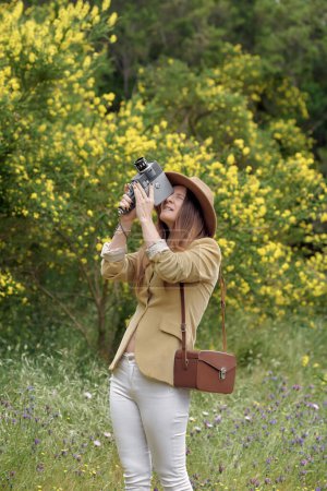 Foto de Fotógrafa en sombrero con ojos cerrados filmando vídeo con cámara de vídeo retro mientras está de pie cerca de árboles con flores amarillas florecientes a la luz del día - Imagen libre de derechos