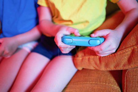 Photo for Cropped unrecognizable siblings with game pads playing video game while sitting on couch in living room with dim light - Royalty Free Image