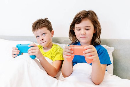 Foto de Hermanos enfocados con gamepads jugando interesante videojuego mientras están sentados juntos en una cómoda cama bajo una manta en el dormitorio en casa - Imagen libre de derechos
