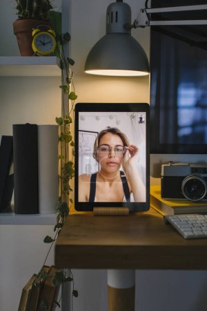 Girl is putting on her glasses during a video call with a friend. The tablet is in a front view over a desk with a computer, some books and a camera.