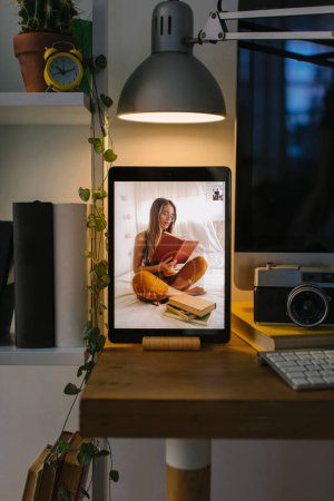 Photo for Girl reading in her bed during a video call in Corona Times. The tablet is in a front view over a desk with a computer, some books and a camera. - Royalty Free Image