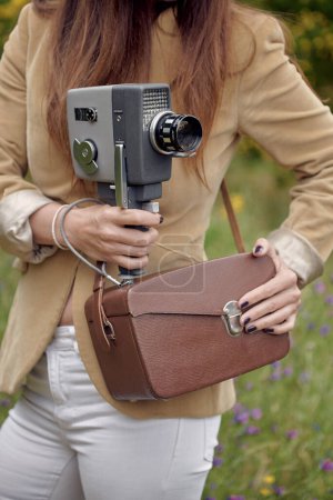 Photo for Crop unrecognizable slim female photographer in jacket standing on grass with leather case and retro video camera in countryside in summer - Royalty Free Image