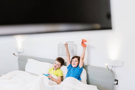 Photo for Boy raising arms with game pad in excitement while playing video game with upset sister in bed in light bedroom - Royalty Free Image