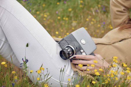 Téléchargez les photos : Vue latérale du photographe anonyme mince en jeans couvrant le visage avec un chapeau tout en étant couché sur la prairie avec des fleurs colorées et tenant une vieille caméra vidéo dans la campagne - en image libre de droit